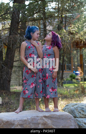 Preteen sisters, in colorful jump suits, in outdoor setting. One has hair dyed blue and the other has red hair, facing each other, standing on rock. M Stock Photo