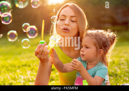 family, happiness, childhood and people concept - Cute female child blows soup foam and make bubbles with her mother in park Stock Photo