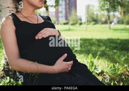 Pregnant woman in black dress sitting on grass in the sunny park Stock Photo