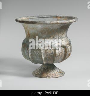 Glass cup with splayed foot. Culture: Roman. Dimensions: H. 5 3/16 in. (13.2 cm); diameter  5 9/16 in. (14.1 cm). Date: 2nd half of 1st century A.D..  Translucent blue green.  Outsplayed tubular rim, folded over and in; tall curving neck, tapering downwards; thick projecting shoulder; squat, globular body; applied conical foot, formed from a separate gather, with edge cracked-off and ground flat; small, circular, flat bottom.  Eleven vertical ribs in high relief tooled out from shoulder down side of body.  Rim and body cracked with one hole in side; part of foot broken and missing; few bubbles Stock Photo
