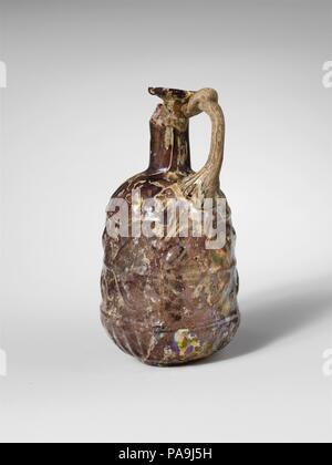 Glass jug. Culture: Roman. Dimensions: H.: 3 1/2 in. (8.9 cm)  Diam.: 1 7/8 in. (4.8 cm). Date: mid-1st century A.D..  Translucent streaky pale purple, with handle in uncertain color.  Rim, folded out, over, and in; broad flaring mouth; slender cylindrical neck; convex curving shoulder; cylindrical body, expanding downwards, then cup-shaped lower section; low circular base, with rounded edge and bottom concave at center; rod handle attached in a pad to shoulder, drawn up, round, and in, and folded onto underside of rim with slight fold above rim. A mold seam runs down sides; a separate cup-sha Stock Photo