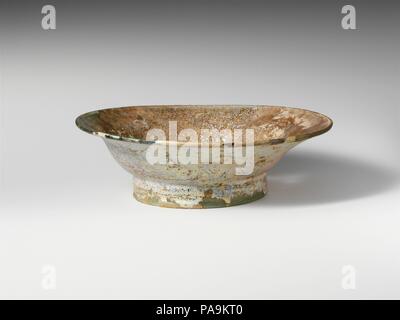 Glass dish. Culture: Roman. Dimensions: H.: 2 5/16 in. (5.9 cm)  Diam.: 7 3/16 x 3 3/4 in. (18.3 x 9.5 cm). Date: 4th century A.D..  Translucent pale blue green.  Thickened, rounded rim; side curves down and in, with angular profile; tall foot ring, made by folding, slightly splayed and with tubular, inward-bulging edge; uneven bottom with thick dome at center and prominent pontil scar.   Intact; many pinprick bubbles; heavily weathered, with pitting, some dulling, and brilliant iridescence. Museum: Metropolitan Museum of Art, New York, USA. Stock Photo