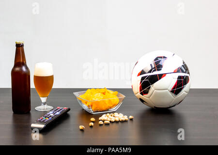 People prepared to watch football on TV with beer. There's beer on the table, ball, TV remote, snacks. Craft beer. Light background. Stock Photo
