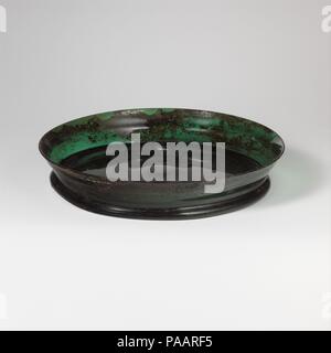 Glass dish. Culture: Roman. Dimensions: H.: 1 5/16 in. (3.3 cm)  Diam.: 6 7/8 in. (17.5 cm). Date: 1st century A.D..  Translucent deep blue green.  Outsplayed, rounded rim; body tapers downwards, with projecting horizontal ridge at mid-point of side; shallower on one side than the other; thick, tubular foot ring at junction with side, made by folding; flat bottom with central pontil scar and slight kick on interior.  Intact; many bubbles; dulling, pitting, and brown, enamel-like, iridescent weathering on exterior, with many surface scratches, little weathering on interior.  Green, shallow, col Stock Photo