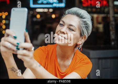 Close up of attractive blonde girl in orange T-shirt making selfie at cafe Stock Photo