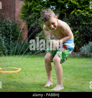 A young 6 year old boy running and playing in the a the garden with water sprinkler. UK Stock Photo