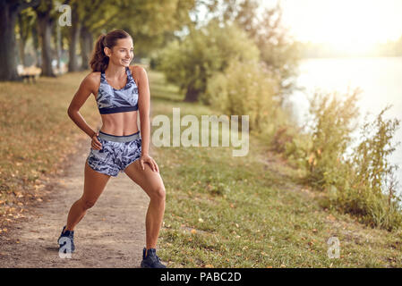 Fit muscular woman working out in a park on a footpath alongside a peaceful lake or river doing stretching exercises in a health and fitness concept Stock Photo