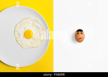 Fried egg on plate with  an uncooked egg  looking worried he's next. Two tone split color background flat lay image. Stock Photo