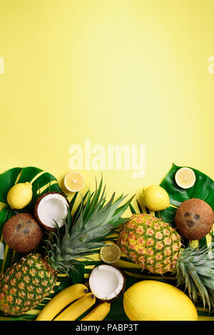 Exotic pineapples, ripe coconuts, banana, melon, lemon, tropical palm and green monstera leaves on yellow background with copyspace. Creative layout.  Stock Photo