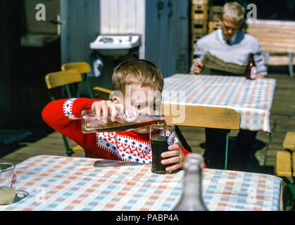 Little boy wearing a Winter patterned jumper pouring Pepsi Cola from an old fashioned bottle into a glass at a cafe table, Hamburg, Germany in the 1960s Stock Photo