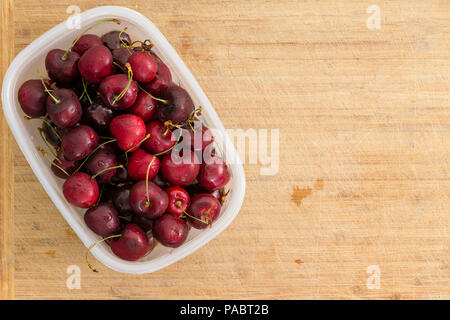 Small plastic container filled with red cherries placed to the side on an old bamboo chopping board ready to serve for a healthy summer dessert rich i Stock Photo