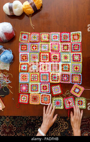 Pastel coloured granny square woollen baby blanket, traditional hand made crochet home handicraft being laid out on a wooden table with balls of wool Stock Photo