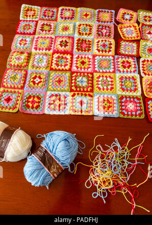 Brightly coloured granny square woollen baby blanket, traditional hand made crochet home handicraft, with balls of blue and white wool and threads Stock Photo