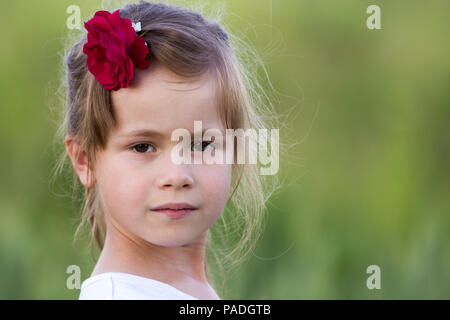 Portrait of beautiful small preschool blond girl with nice gray eyes and red rose in hair smiling dreamily in camera on blurred bright green backgroun Stock Photo