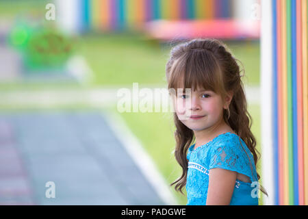 Portrait of little fashionable blond girl in blue dress, with gray eyes and beautiful long hair smiling in camera on bright blurred colorful backgroun Stock Photo