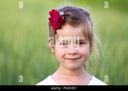 Portrait of beautiful small preschool blond girl with nice gray eyes and red rose in hair smiling dreamily in camera on blurred bright green backgroun Stock Photo