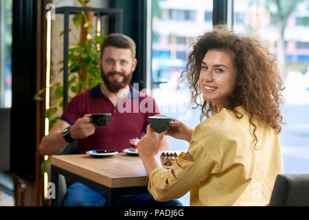 Smiling couple having date in restaurant, keeping coffee cups, looking at camera, sitting on small wooden table near big, panoramic windows. Wearing colorful casual stylish clothes