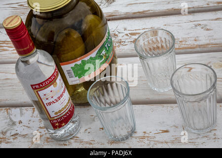 A bottle the of vodka 'Festive', three faceted glasses and a can of salted cucumbers lie on a wooden table Stock Photo