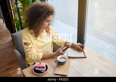 Pretty curly girl writing notes, drinking coffee in caffee. eating cake, studying, sitting on wooden table near big window. Wearing yellow blouse, having pretty face, big eyes, plump lips. Stock Photo