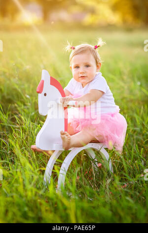 beautiful little girl in pink dress riding on wooden toy horse outdoors Stock Photo