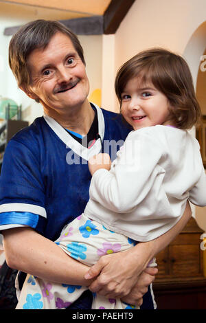 A man with Downs Syndrome carrying his great niece.  They are both happy and looking at the camera.  She is wearing pajamas and he has at least three  Stock Photo