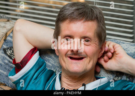 A happy man with Down Syndrome leans back on a couch with a big smile.  He is older, and has lost all his teeth.  He is looking at the camera and has  Stock Photo