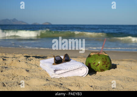 Sunglasses, towel and a fresh young coconut on the sandy beach with blurred Atlantic ocean in background, Rio de Janeiro, Brazil Stock Photo