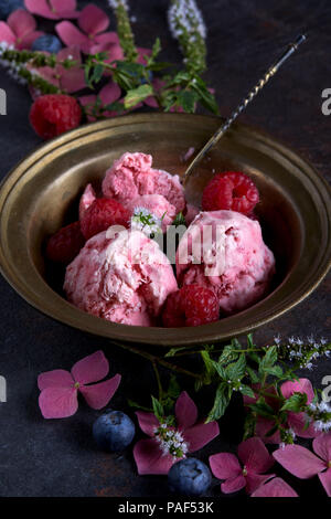 Homemade raspberry ice cream in copper bowl on table with berries and flowers from the garden. Stock Photo