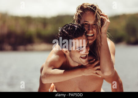 Portrait of happy young man piggybacking his girlfriend. Cheerful young couple enjoying their vacation on lakeside. Stock Photo