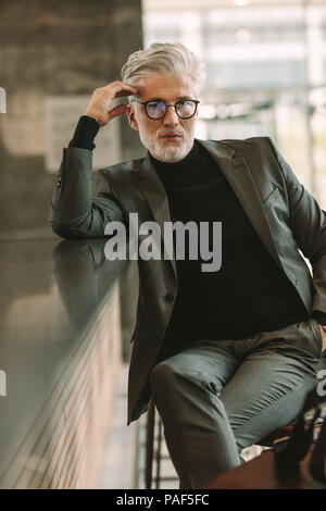 Vertical portrait of handsome mature businessman sitting at cafe counter and looking at camera. Senior man in business suit sitting at coffee shop. Stock Photo