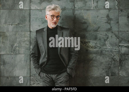 Portrait of handsome mature man in business suit standing on grey wall. Stylish businessman standing with hands in pocket and looking at camera. Stock Photo
