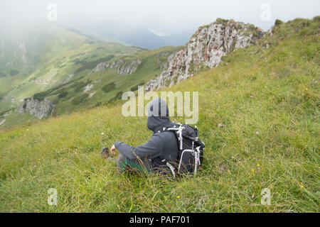 rear view of hiker with backpack sitting on green grass in mountain landscape enjoying scenic view Stock Photo