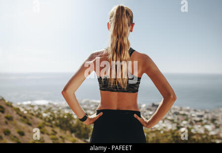 Rear view of young woman's standing with her hands on hips and looking at a view. Female runner taking break from running. Stock Photo