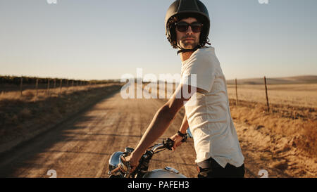 Young man wearing a helmet and sunglasses standing with his bike and looking back on rural road. Male bike on countryside road. Stock Photo