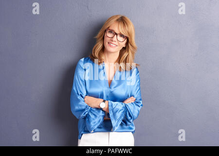 Portrait of an attractive middle aged woman with toothy smile and arms crossed posing against a grey background in the studio. Stock Photo
