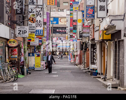 Japanese businessman with briefcase walks towards camera long Tokyo backstreet with crowded walls covered in signs for shops Stock Photo