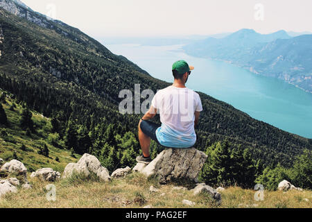 Young man sitting on the hill top and enjoying valley view. Succes, vacation and travel concept. Monte Baldo mountain, Lago di Garda lake, Veneto region, Italy. Stock Photo