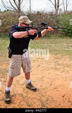 A man stands in a wooded area looking through the scope of an AR-15 rifle while aiming Stock Photo