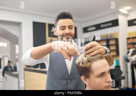 Smiling hairstylist doing a hairdress for young client in beaty salon. Barber using sharp metallic scissors, wearing grey waistcoast, white classic shirt. Care products for hair on background. Stock Photo