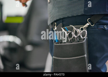 Close up of barber's leather bag with metallic sharp scissors hanging on waist. Hairstyler wearing dark blue trousers, grey waistcoat. Going to work with client waiting for new hairdress. Stock Photo