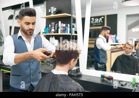 Doing new modern hairstyle for young man in barber shop. Professional hairstylist using clipper, looking concentrated. Male model sitting in front of big mirror, covered with special black cape. Stock Photo