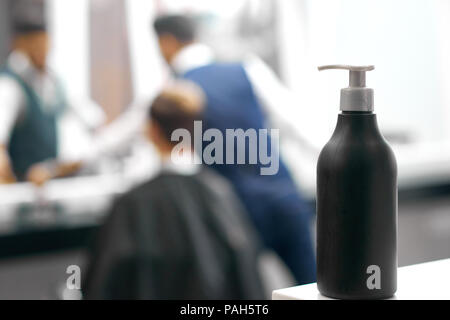 Photo of black plastic bottle with dispenser including hair care product. Standing on white glance surface in barber shop, beaty salon. Hairdresser working with client near mirror on background. Stock Photo