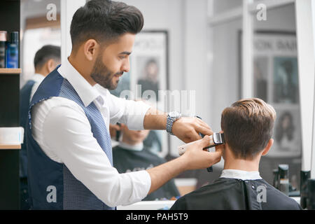 Barber doing new haircut for young client sitting in front of mirror. Wearing white casual shirt, grey waistcoat, watch. Looking concentrated, loving his job. Model covered with special black cape. Stock Photo