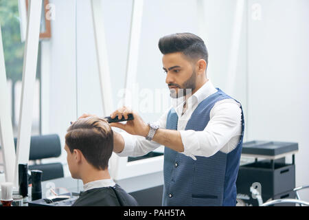 Barber doing new haircut for young client using black plastic comb. Wearing white casual shirt, grey waistcoat, watch. Looking concentrated, loving his job. Model covered with special black cape. Stock Photo