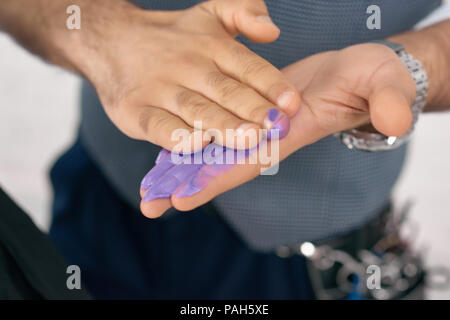Haircolorist's hands smeared with paint spots. Hairdresser doing hair coloring with violet, lilac tint. Wearing stylish man's handwatch. Stylish waistcoat on photo's background. Soft focus.