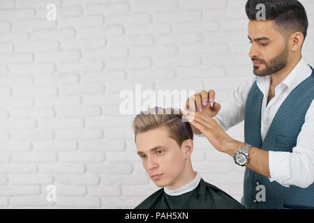 Barber doing lilac hair coloring for young handsome client. Hairdresser wearing grey waistcoat, white shirt, watch. Boy's clothes covered with special black cape. Sitting on white studio background.