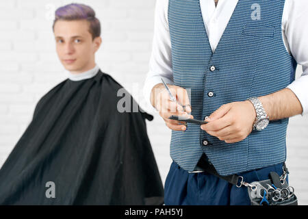 Cropped photo of barber keeping black plastic comb and scissors istanding in front ofyoung client. Boy having toned in violet color hair and looking at camera. Master wearing scissors case, waistcoat.