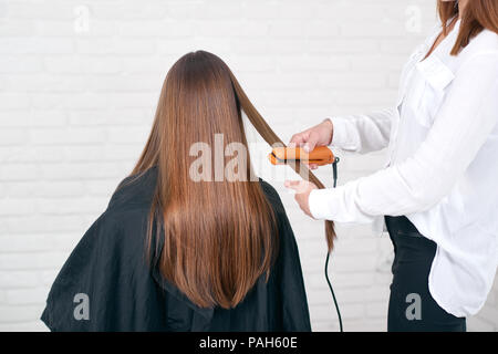 Model sitting while hairstyling in beaty salon with white brick walls. Wearing black cape for hairdressing. Girl having dark, long, glancy, straight, brown hair. Hairdresser using hair flatter. Stock Photo