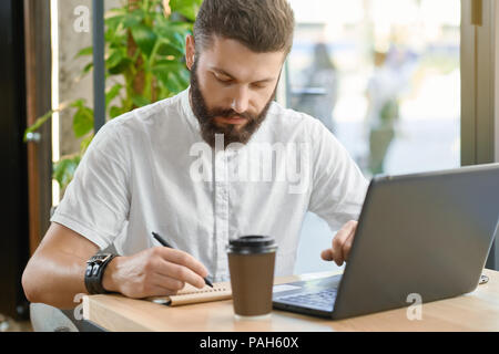 Bearded man writing notes, working with laptop sitting near window in restaurant. Drinking coffee fron paper can, keeping a pen. Wearing white casual shirt, leather bracelet. Stylish hipster. Stock Photo