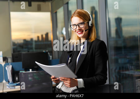 Photo of smiling woman with glasses and headphones with paper in hands near glass wall Stock Photo